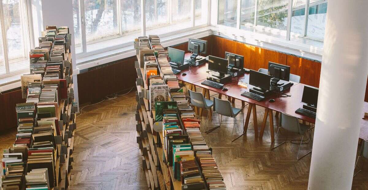 Conduct school facility assessment | A beautiful library with many books stacked neatly on two long shelves, and some desktops on a table