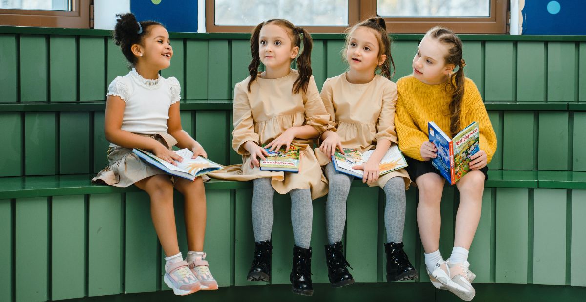 Four young female children smiling as they read their books on a green bench in their classroom